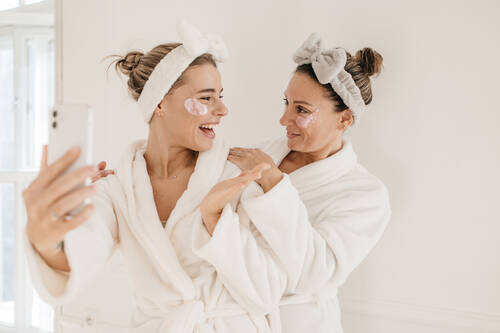 Happy young caucasian women wear bathrobes use modern smartphone white background home spa concept